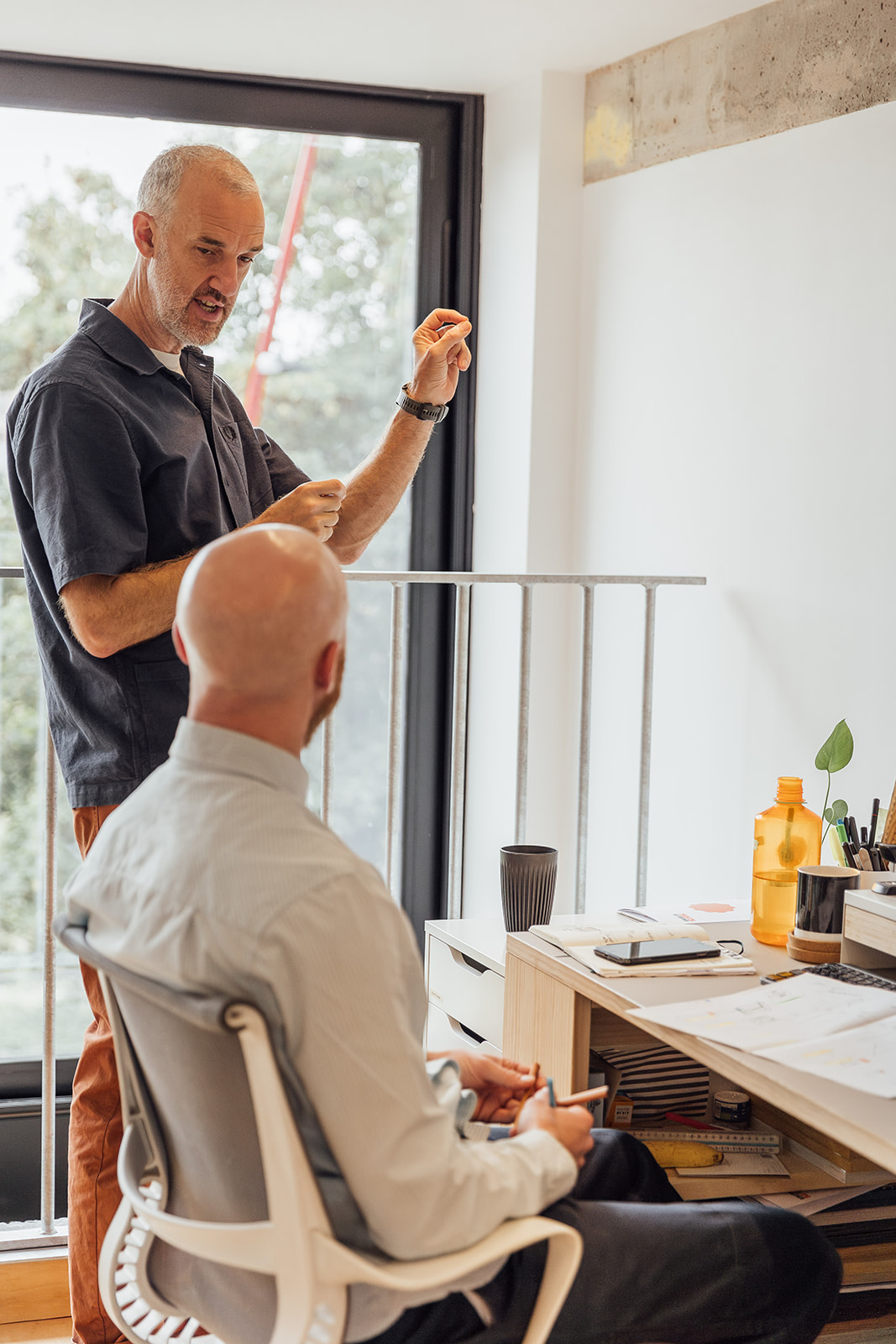 A man with a grey shirt directing a man sitting at their desk